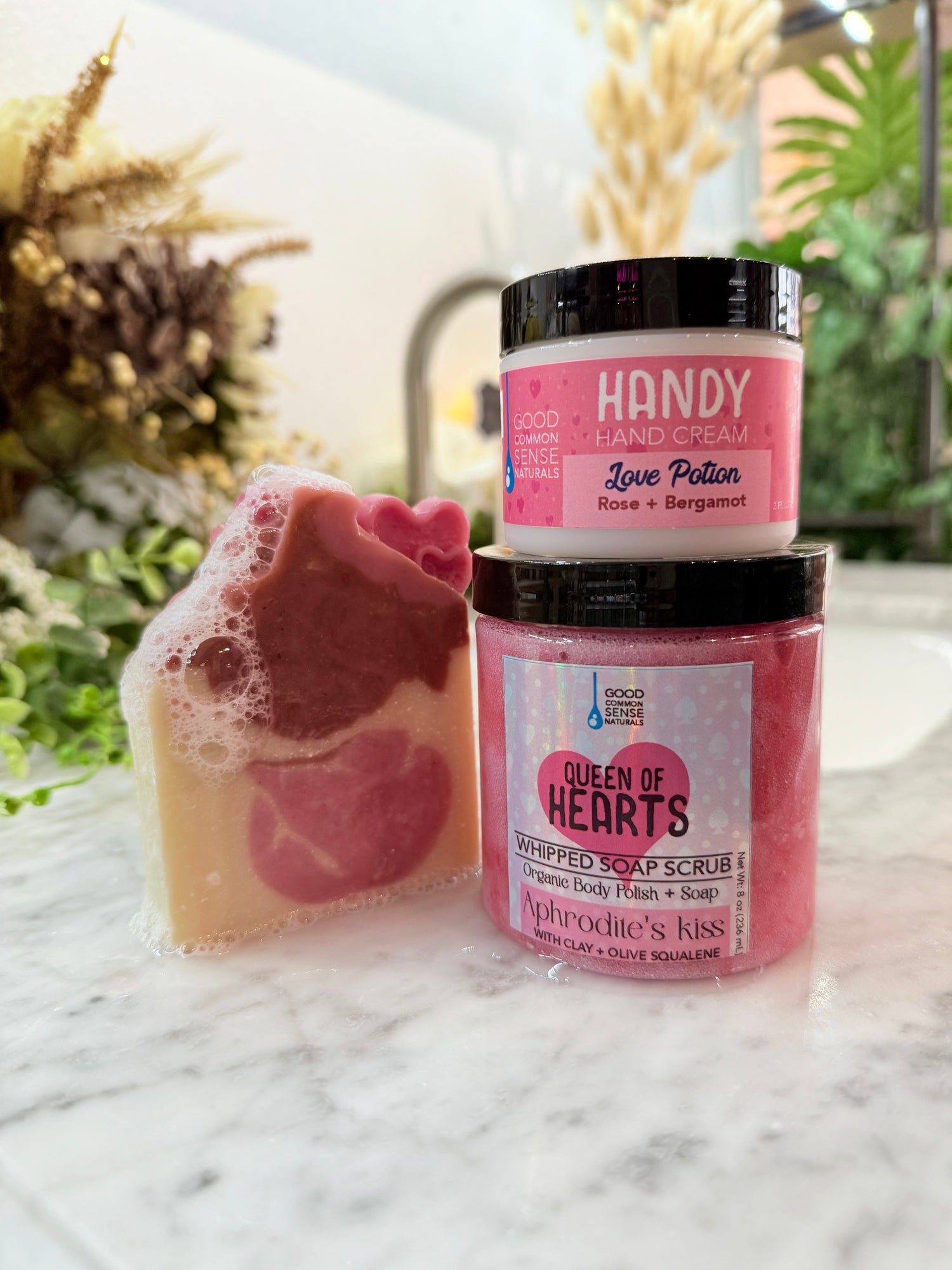 Handcrafted pink heart-shaped soap displayed beside Queen of Hearts body scrub and Love Potion Handy Cream on a marble counter
