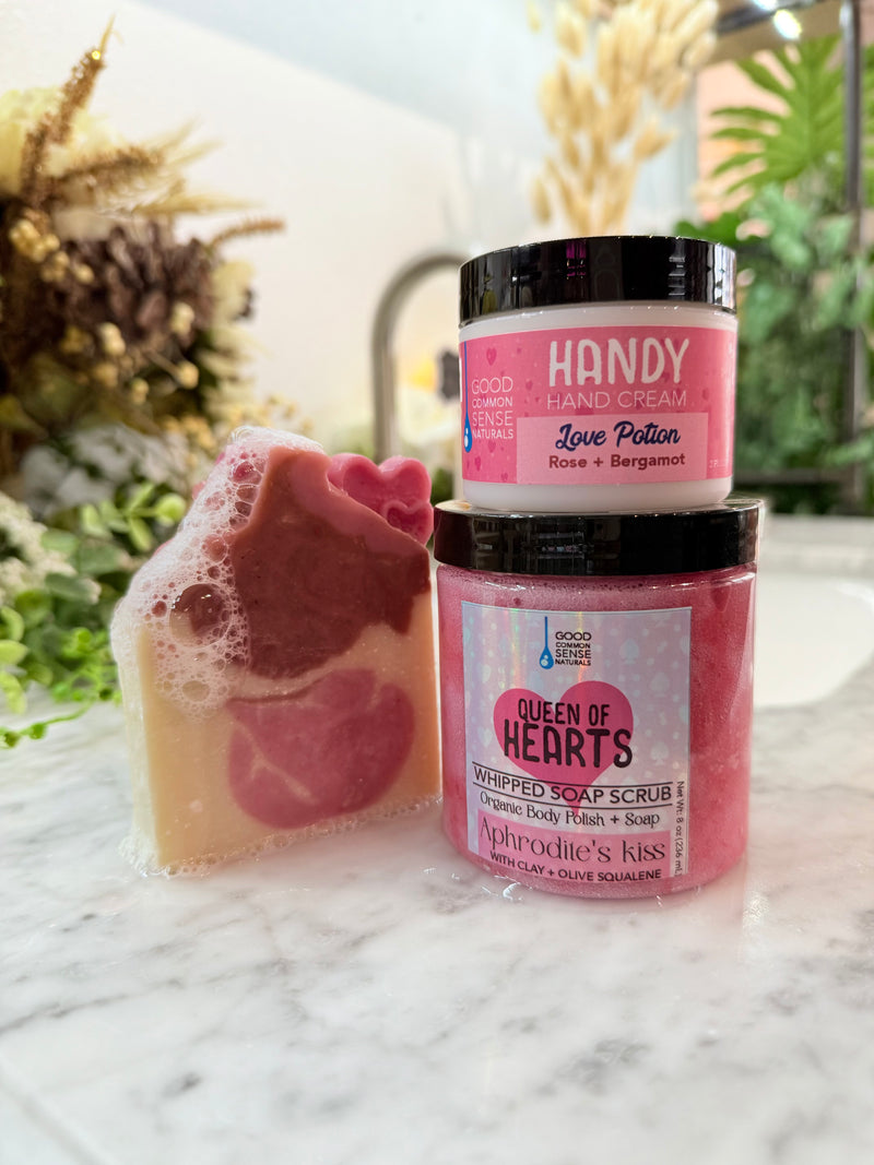 Handcrafted pink heart-shaped soap displayed beside Queen of Hearts body scrub and Love Potion Handy Cream on a marble counter