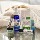 A collection of skincare products including a spray bottle, a deodorant stick, lip balm, hand cream, moisturizer, and aromatherapy roller, arranged on a reflective surface with a blurred background.