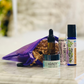 A travel gift set with a purple zippered pouch containing a nail serum, hand cream, lip balm, and essential oil blend, displayed on a marble surface.