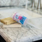 A bottle of "I Can't Adult Today, Bubble Push Pop" lies on its side with a Bubble Pop wand on a wooden tray, on top a marble countertop near a sink.