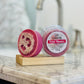 A round, pink I Loofah You Petal Meadow Soap rests on a wooden soap holder atop a marble countertop, with a blurred bathroom sink and faucet in the background.