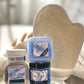 A gift set containing various herbal skin care products, including a scrubbing body mitt, sugar scrub, mini buttah, salt soak, and lotion bar, displayed on a soft surface.