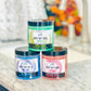 Three jars of Buff My Soul sugar scrub in blue, green, and red colors, placed on a marble surface.