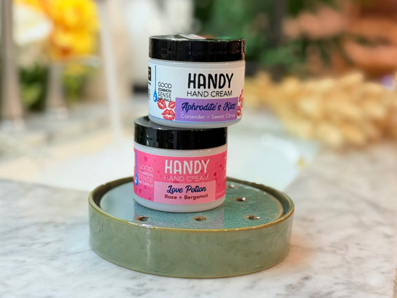 Two jars of HANDY hand cream stacked on a decorative tray with a blurred background