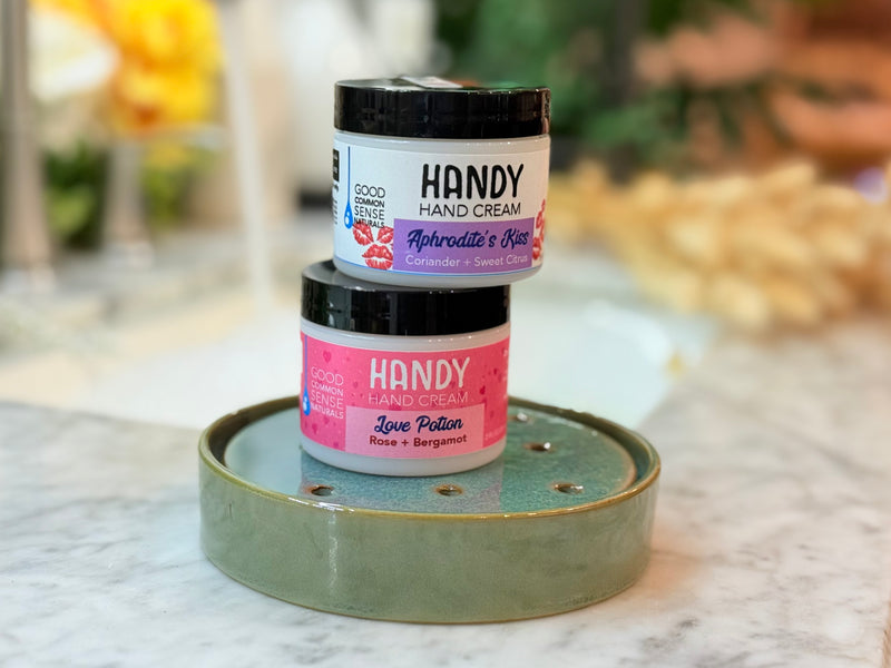 Two jars of HANDY hand cream stacked on a decorative tray with a blurred background