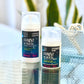 Two bottles of 'Fairest of Them All' facial serum, one smaller and one larger, placed on a reflective surface with a blurred background featuring plants.