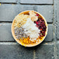 A wooden bowl filled with various dried herbal bath additives such as calendula, marshmallow root, coconut milk, slippery elm, hibiscus, and lavender, placed on a dark tiled surface.