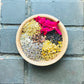 A wooden bowl filled with a mixture of dried herbs including chamomile, lavender, white sage, yarrow, elderberry, clove, and rose, placed on a dark tiled surface.