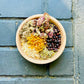 A bowl filled with various dried herbs, including oatmeal, mugwort, calendula, echinacea, juniper, and red clover, arranged for a ritual bath kit on a dark tiled surface.