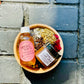 brown bowl filled with herbs with a body oil and sugar scrub set on top. with a black bricked backround  