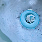 A close-up image of a bath bomb with a blue bubbly frosting design, resembling a donut, in a bath of bubbles.