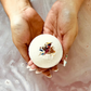 A person's hands holding a bath bomb over water with a visible sprinkle of rose petals on top.