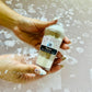 A person holding a small, plastic frap cup with a bath bomb mixture inside with a mocha color scheme, with bubbles rising around their hand in a bubbling bath.