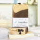 A bar of handmade soap with a white and brown color scheme, labeled 'Twisted Bean', positioned on a wooden dish with a blurred background.