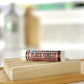 A tube of Plaid Forest lip balm resting on a wooden surface with a blurred background.