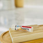 A tube of 'Immortal' lip balm on a wooden dish, indicating a beige color 
