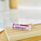 A tube of Heart Beet tinted lip balm placed on a wooden dish with a blurred background.