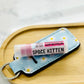 A tube of pink lipstick labeled 'Space Kitten' with a blue and white patterned case, resting on a bamboo makeup tray.