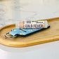 A tube of Gold Fever lipstick shimmer is placed on a wooden tray next to a blue keychain, on a marble surface.