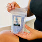 A person holding a small clear plastic bag with blue bath salts and a label that reads 'Float Away, Lavender Myrrh'. The bag is being held in front of a bathtub.