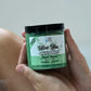 A container of Olive You Sugar Body Scrub - Sauna Rescue held in a person's hand, with a green label and clear contents visible.
