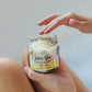 A person's hand holding an open jar of sugar scrub with another hand reaching for it, with the label 'Olive You Sugar Scrub - High Pine' visible.