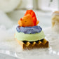 green bath bomb on a wooden soap dish with a whipped blue body butter and an orange bath toy on top