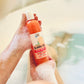 A person holds a bottle of Bubbles Up! Simply Joyful Body Wash with an orange label, surrounded by bubbles in the tub—legs partly visible