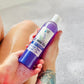 A tattooed person holds a bottle of Bubbles Up! Lavender Plains Body Wash in a bubble bath, their legs partially visible and soap suds on their hand.