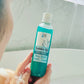 A person in a bathtub holds a blue-green bottle of Bubbles Up! Sauna Rescue Body Wash with a white label featuring eucalyptus leaves. Tiled bathroom walls and a sunlit window appear in the background.