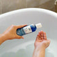 A person is using a blue bottle of Silkening Cream body wash in a bathtub.