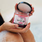 A person holds an open jar of Buff My Soul Pink emulsified sugar scrub with calendula extract in their wet, soapy hands. The label shows the scent is sweet vanilla.