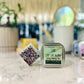 A Cooling Lotion Bar - Ice Ice Baby sits beside an open tin, with a blurred backdrop of indoor plants and home décor.