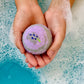 Two hands hold the Lavender Lumberyard Bath Bomb, featuring a small lavender flower design, above blue, bubbly bathwater—perfect for creating a luxurious bath experience.