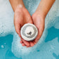 A person holding a bath bomb in their hands, with the bath bomb partially submerged in bubbles with a blurry background.