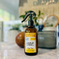 A brown spray bottle with a black spray nozzle labeled 'Lounge High Pine, Room & Linen Spray' placed on a marble surface with a plant and decorative items in the background.