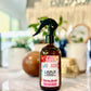 A bottle of Lounge Jasmine Gardens room and linen spray with a spray nozzle, placed on a surface. The label has floral graphics and the scent marked as Jasmine & Chamomile.