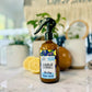 A brown spray bottle labeled 'Lounge' with a green spray nozzle, placed on a light surface with a plant and decorative items in the background.