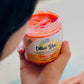 Someone holds an open jar of Olive You Sugar Scrub - Simply Joyful, a bright orange body scrub made with organic sugar and olive squalene. It has an orange and vanilla scent and a label with cheerful citrus illustrations.