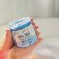 A person holds a jar of Olive You Sugar Scrub - Signature Spa, filled with visible blue scrub. The label notes its lavender, sage, and eucalyptus scents. The background is gently blurred.