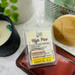 A wax melt in a clear container with a label that reads 'High Pine Manzanillo Wax Melts'. placed on a marble surface with a plant and lotion  candle in backround 