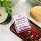 A package of Jasmine Tea Wax Melts by Good Common Sense, placed in a brown wax melt container, with a label that includes the product name and descriptions, placed on a marble surface with a few dried jasmine flowers and green tea leaves in the background.