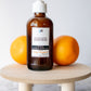 A bottle of olive oil hand soap on a wooden dish with two whole oranges and one half-cut orange beside it, placed against a white background.
