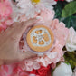 A hand holding a bar of fruity loopy exfoliating soap, with a decorative design on the top, surrounded by pink and red flowers.