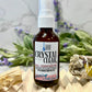 A bottle of 'Crystal Clear' aura spray with a label that includes the product name and a brief description, displayed on a wooden surface with a background of flowers and a crystal.