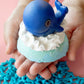 blue bath bomb topped with a white whipped body cream and a toy whale on top on the hands of a person 