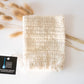 A beige soap saver pouch made of sisal with a drawstring, placed on a white surface, flanked by two decorative dry plants.