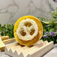 A bar of handcrafted soap with a textured, sponge-like appearance, placed on a wooden dish with a marble-like background and some purple flowers in the foreground.