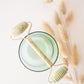 A jade roller with a green glass base, gold-colored handle, and a pampas grass head resting on a white surface.